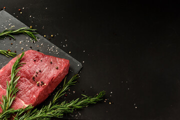 piece of beef with rosemary, pepper and salt on a dark background with copy space.