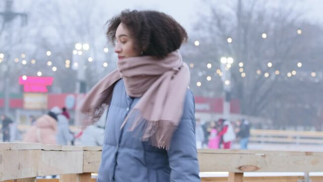 Beautiful positive afro american girl ethnic millennial girl stands outdoors against background of city ice rink people skating, lady teenager with curls dances enjoying moves celebrating weekend