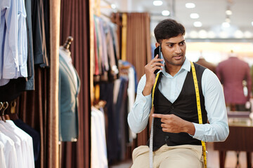 Portrait of serious young Indian tailor working in fashion atelier and talking on phone with client