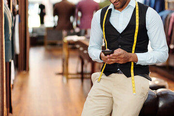 Cropped image of young tailor leaning on chair and checking text messages in smartphone during break