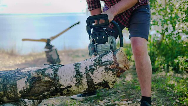 A woodcutter, logger saws a firewood with a chainsaw on the beach of a lake, river. Two axes are stuck in a stump in the background. High quality 4k footage