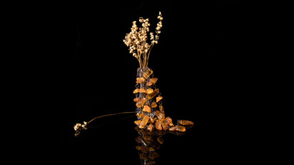 Retro composition of Baltic amber necklace on a glass bottle with dried lilies of the valley on a black background. 