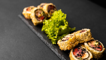 Custom sushi roll in tempura with nori, fresh salmon, tuna, avocado, masago caviar, drizzled with pineapple sauce with salad pouring as decoration on a black plate on a black table and background.