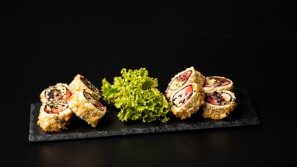 Custom sushi roll in tempura with nori, fresh salmon, tuna, avocado, masago caviar, drizzled with pineapple sauce with salad pouring as decoration on a black plate on a black table and background.