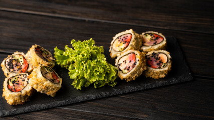 Custom sushi roll in tempura with nori, fresh salmon, tuna, avocado, masago caviar, drizzled with pineapple sauce with salad pouring as decoration on a black plate on a wooden table and background.