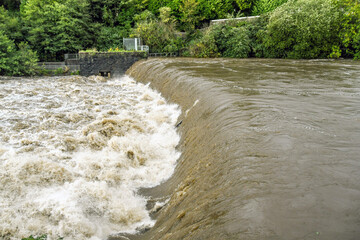 Turbulent water at a weir of a river in flood after extreme weather. No people. - obrazy, fototapety, plakaty