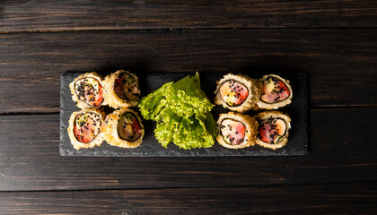 Custom sushi roll in tempura with nori, fresh salmon, tuna, avocado, masago caviar, drizzled with pineapple sauce with salad pouring as decoration on a black plate on a wooden table and background.