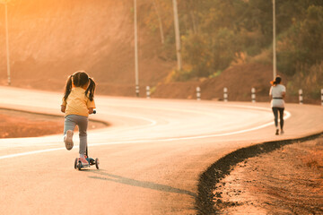 Mother and child girl jogging with stroller road rollers skateboard on countryside road at sunset light