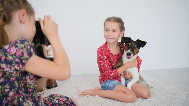 Little girl taking pictures with film camera of sister with fox terrier dog sitting on floor in light room