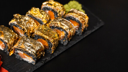 Custom sushi roll with nori, cuttlefish ink, fresh salmon, cream cheese, pepper pumpkin, eel, unagi sauce and edible gold leaf with red ginger, wasabi on a square plate on black table and background.