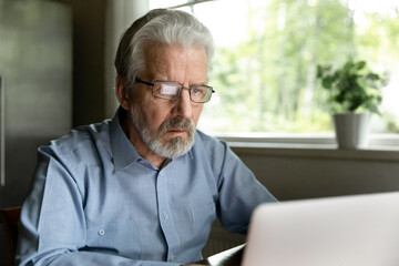 Close up focused mature man wearing glasses using laptop, looking at computer screen, searching information, watching video, chatting with relatives in social network, elderly freelancer working
