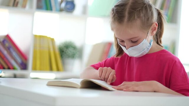 Little schoolgirl in protective mask reading book in classroom