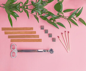 Flat lay of wooden wicks, scissors and wick holders on pink background. Nearby are matches and a branch with green juicy leaves.