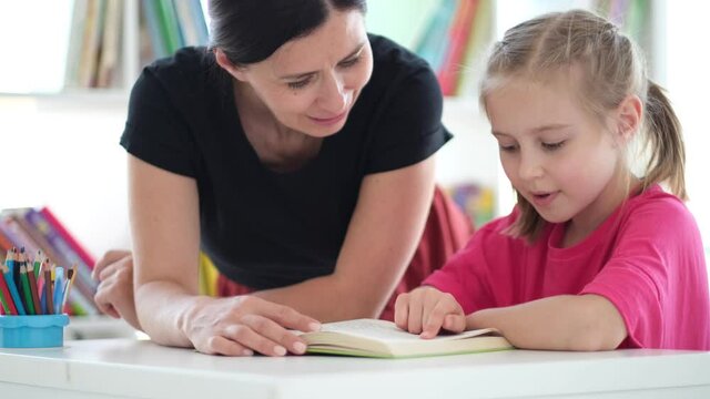 Little girl reading book with help of teacher in classroom
