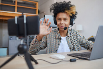 Portrait of teenage African-American boy wearing headset and waving at camera while streaming video games at home, young gamer or blogger concept, copy space