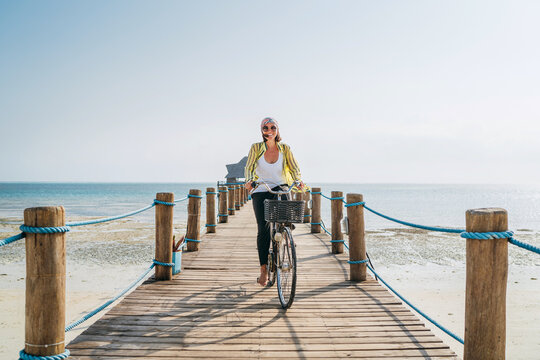Portrait of a happy smiling woman dressed in light summer clothes and sunglasses riding a bicycle on the wooden sea pier and looking at camera. Careless vacation in tropical countries concept image