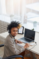 Vertical portrait of teenage African-American boy wearing headset and looking at camera while using laptop at home, young gamer or blogger concept, copy space