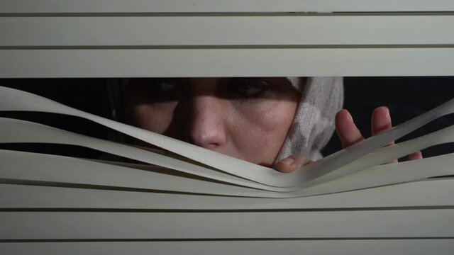 Close up front portrait from indoors of brunette woman in headscarf pushing blinds at window with hands at night. Looking fearfully outside