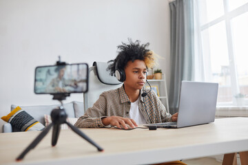 Portrait of teenage African-American boy wearing headset and using laptop while streaming video games at home, young gamer or blogger concept, copy space