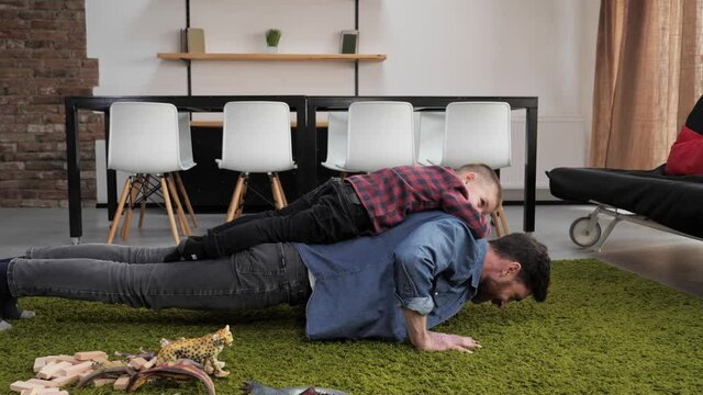 Small cute son playing with his dad in the living room doing a piggy back ride smiling dad doing push ups exercise playful mood having good time together. Father son time. Parents and children.