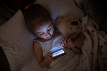 A cute little girl lies on a pillow and enthusiastically plays on the phone at night in bed instead...