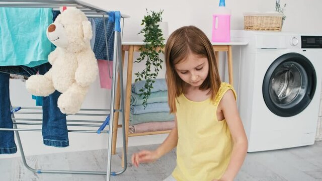 Little girl hanging wet teddy bear toys on dryer using clothespins in room with washing machine