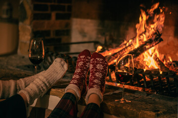 Cosy winter evening. Drinking wine and reading book next to fireplace. Feets in woollen socks by the Christmas fireplace - obrazy, fototapety, plakaty