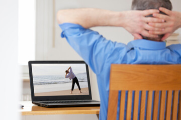 Businessman in home office doing stretching or relaxation exercise in front of laptop with man doing yoga on a beach