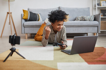 Portrait of emotional African-American boy playing videogames while lying on floor at home with camera filming for online streaming, copy space © Seventyfour