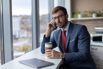Portrait of young handsome man in eyeglasses talking on the phone at his coffee break in the office...