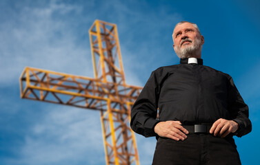 A priest in a black shirt and white clerical collar stands in front of the summit cross on Mount...