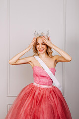 Surprised happy young blonde woman wearing a crown, pink dress and a white ribbon, smiling. Beauty contest winner concept.