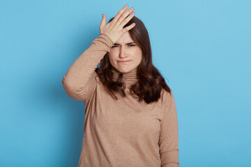 Disappointed woman keeps palm on forehead, regrets, did something wrong, having problematic situation, dressed in casual outfit, poses on blue wall, forgets about important task, lady with pursed lips