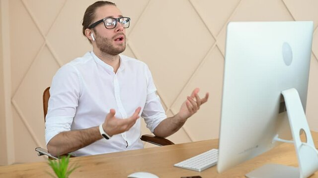 Smiling Male Office Employee Has Pleasant Video Meeting, Virtual Conference With A Team, Confident Hipster Wearing White Shirt And Glasses Looks To PC Webcam And Talking At Ease