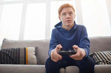 Low angle portrait of red haired teenage boy playing video games while sitting on sofa and holding gamepad, copy space