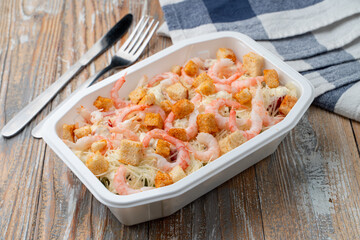 Food delivery: seafood salad with shrimps and croutons in a plastic box on light wooden table