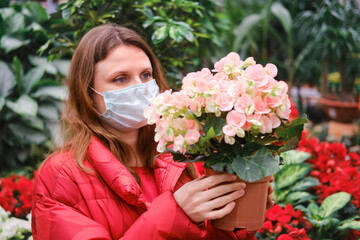 Young woman florist in medical mask chooses begonia flowers in shop. Home gardening hobbies and growing flowers during corona virus isolation