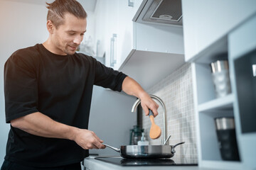 smiling attractive man cooking in pan with wooden spoon at modern home kitchen