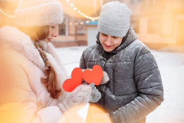 Young loving couple outside on a winter day holding red paper hearts in their hands feeling joy and love. Romantic man and woman celebrate Valentine's day under the snow at a park. Holidays concept.