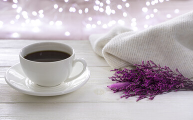 A purple bouquet of flowers, a white cup of coffee and a sweater on the background of a burning garland. Side view with copy space. The concept of holiday backgrounds.
