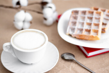 Viennese waffles on a white plate and a cup of cappuccino coffee laid out next to a branch of cotton