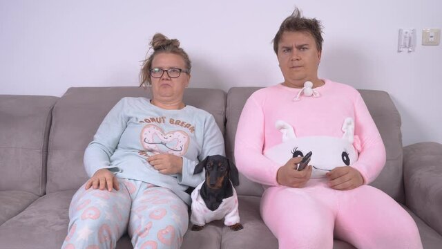 Young dismayed woman and man wearing funny pajamas with adorable dachshund puppy watch TV on comfortable sofa in living room