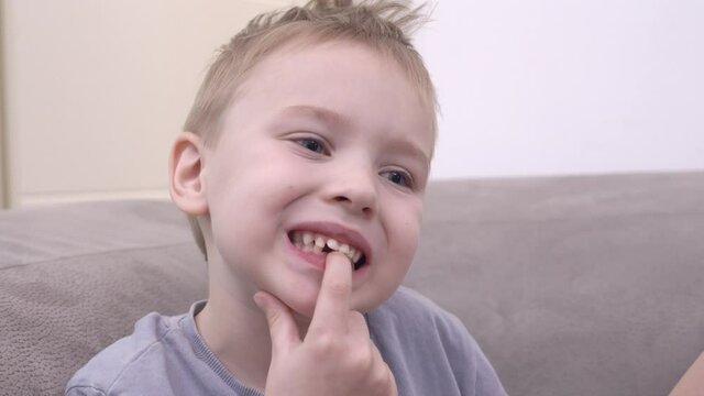 Funny little boy in t-shirt loosens milk tooth on lower jawbone sitting on comfortable sofa in light living room close view