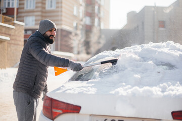A young man cleans his car after a snowfall on a sunny, frosty day.