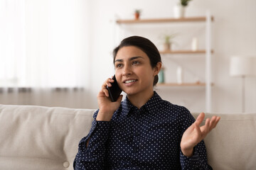 Emotional millennial mixed race female sit on couch talk chat discuss pleasant thing with friend holding mobile phone at ear. Young indian female caller relax at home enjoy good telephone conversation