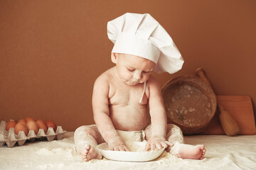 Little boy baker in a chef's hat sitting on the table playing with flour on a brown background with a wooden rolling pin, a round rustic sieve and eggs. Copy, empty space for text