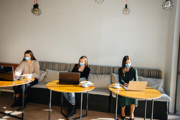 A group of masked girls keep a social distance in a cafe when working on laptops.