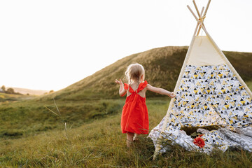 cute little girl is having fun near wigwam in a summer field on sunset. Young family spending time together on vacation, outdoors. The concept of summer holiday. © Andriy Medvediuk