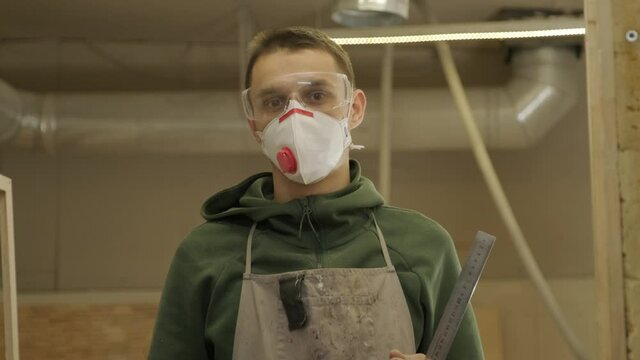 Portrait of worker carpenter with respirator and glasses in workshop, holds the ruler