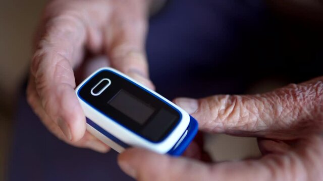 Senior adult with wrinkled hands holding digital pulse oximeter and looking at device, old sick man using oxymeter for measuring oxygen rate in blood. Cardiology problems of elderly man and monitoring